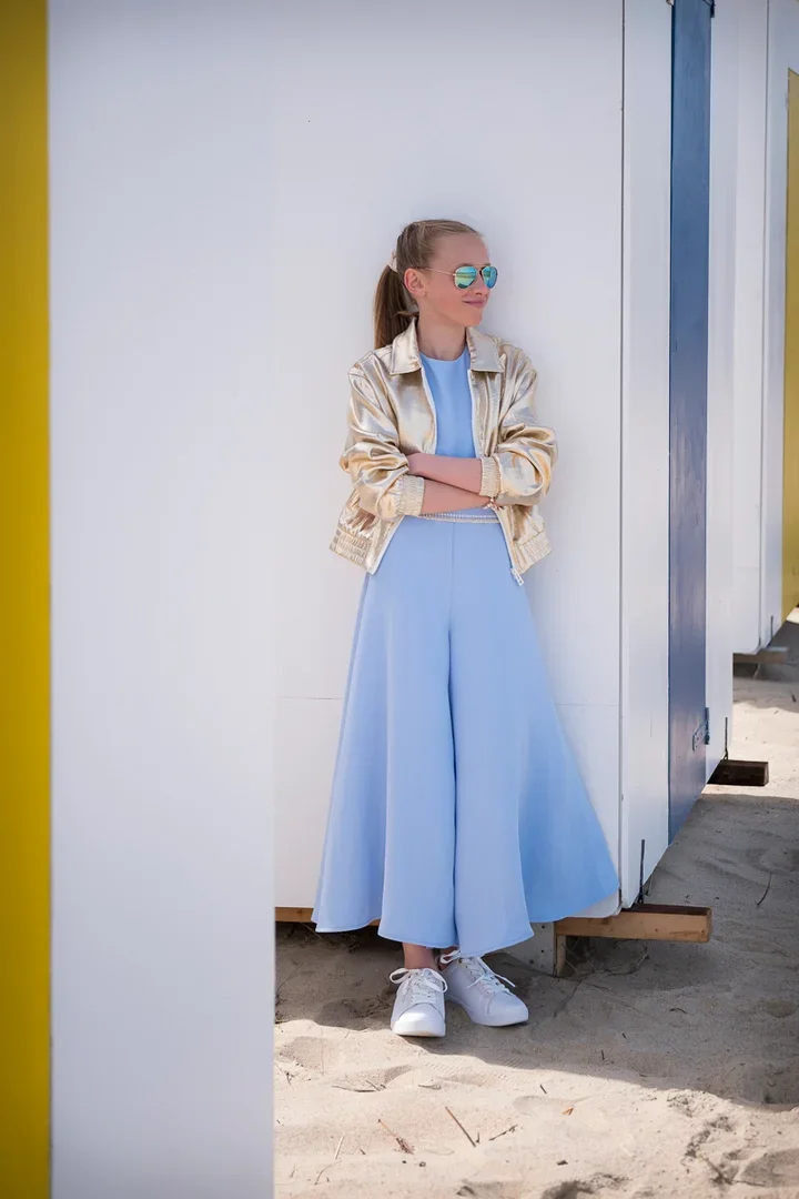 Een tienermeisje met goudkleurige jack, blauwe soepel vallende jumpsuit, witte sneakers en blauw-zilveren zonnebril staat tegen een witte muur op het strand, lachend en met armen gekruist.