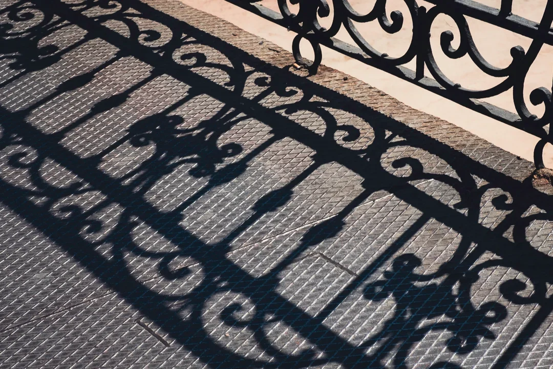 Schaduw van een smeedijzeren hek op een betegeld trottoir.