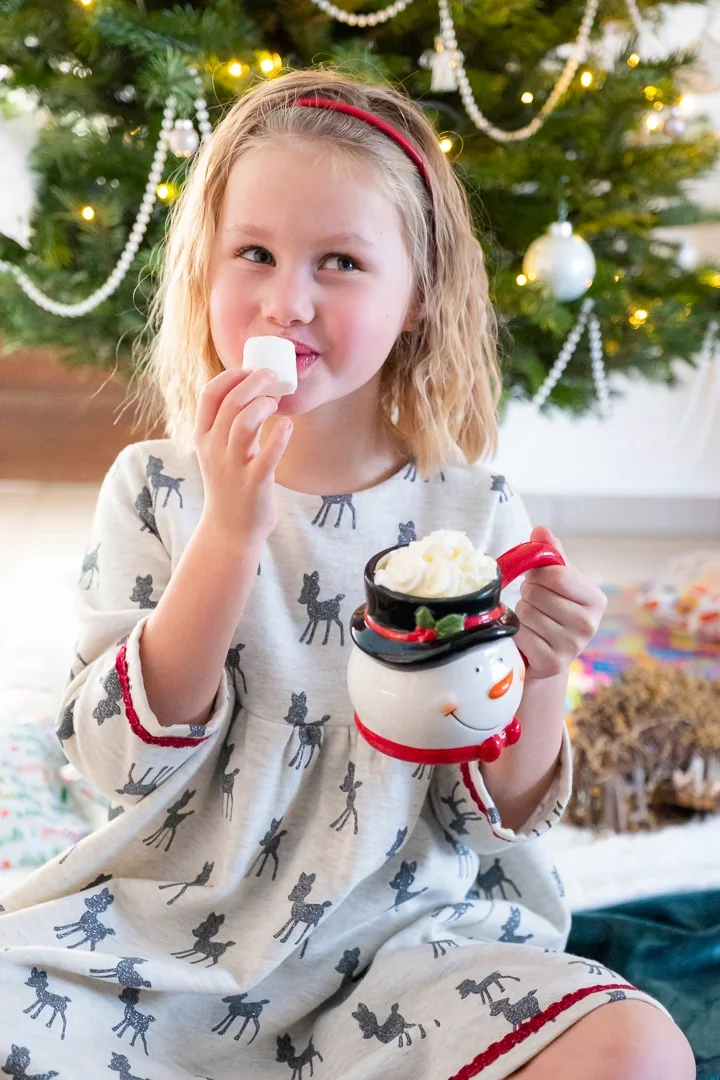 Een jong meisje met blond, golvend haar en een rode haarband, zit voor een versierde kerstboom, en eet een marshmallow terwijl ze een kerstmok vasthoudt.