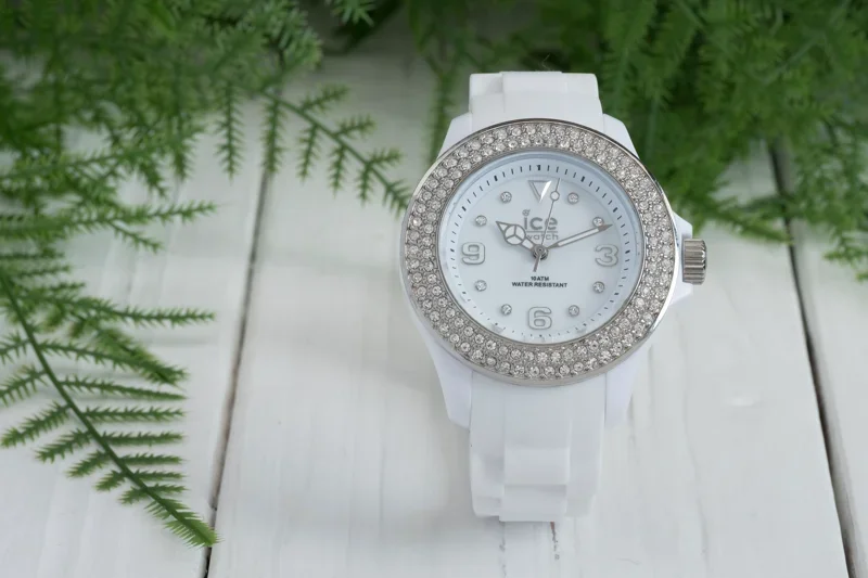 Witte polshorloge met een diamanten versiering op de wijzerplaat, ligt op een witte houten ondergrond met groene planten ernaast.