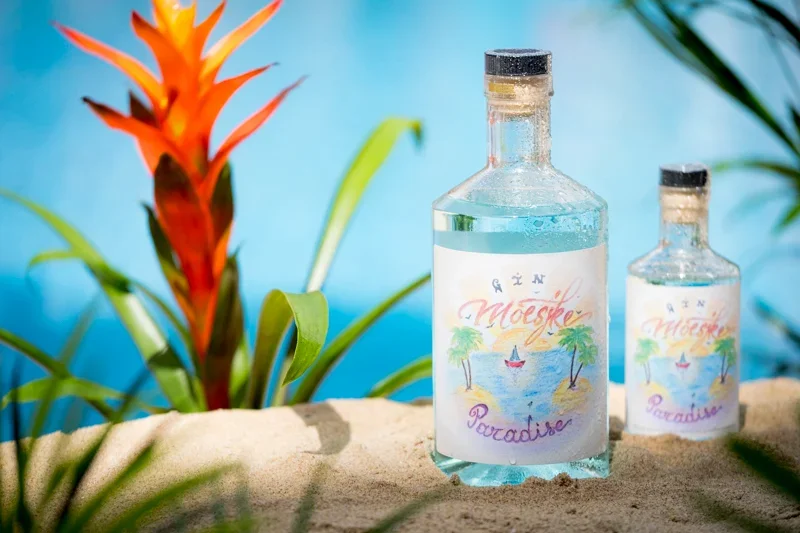 Fotografie van twee glazen flessen met tropisch design op zand, met groene planten en een bloemenstruik op de achtergrond, tegen een blauwe muur.