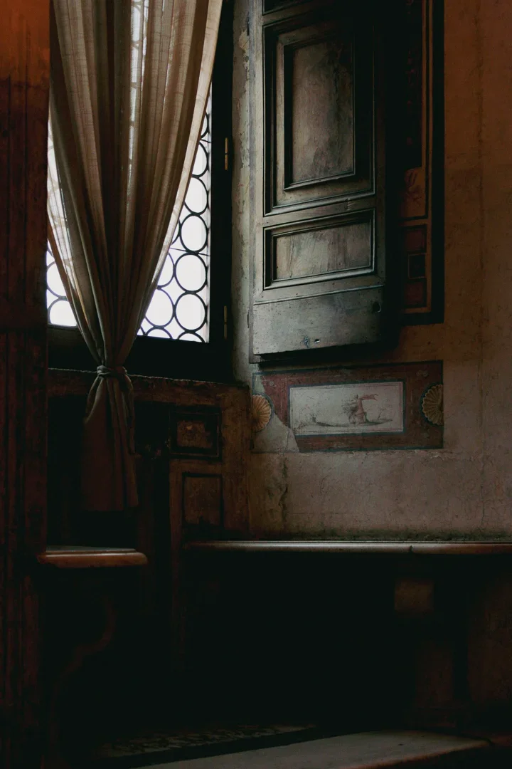 Een kamer met een open houten raam met een ijzeren rooster, een beige gordijn die naar binnen valt, en verlichte muur met oude fresco's.