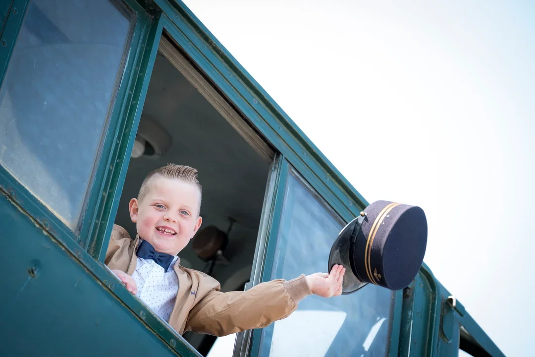 Lachende jongen steekt hoof en arm uit treinraam, zwaait met condecteurshoed.