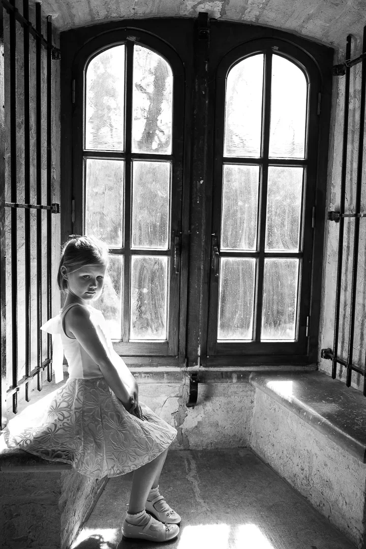 Een jonge meisje zit op een bankje in een kamer met grote, oude ramen en metalen tralies, mogelijks kasteel. Het is zwart-wit.