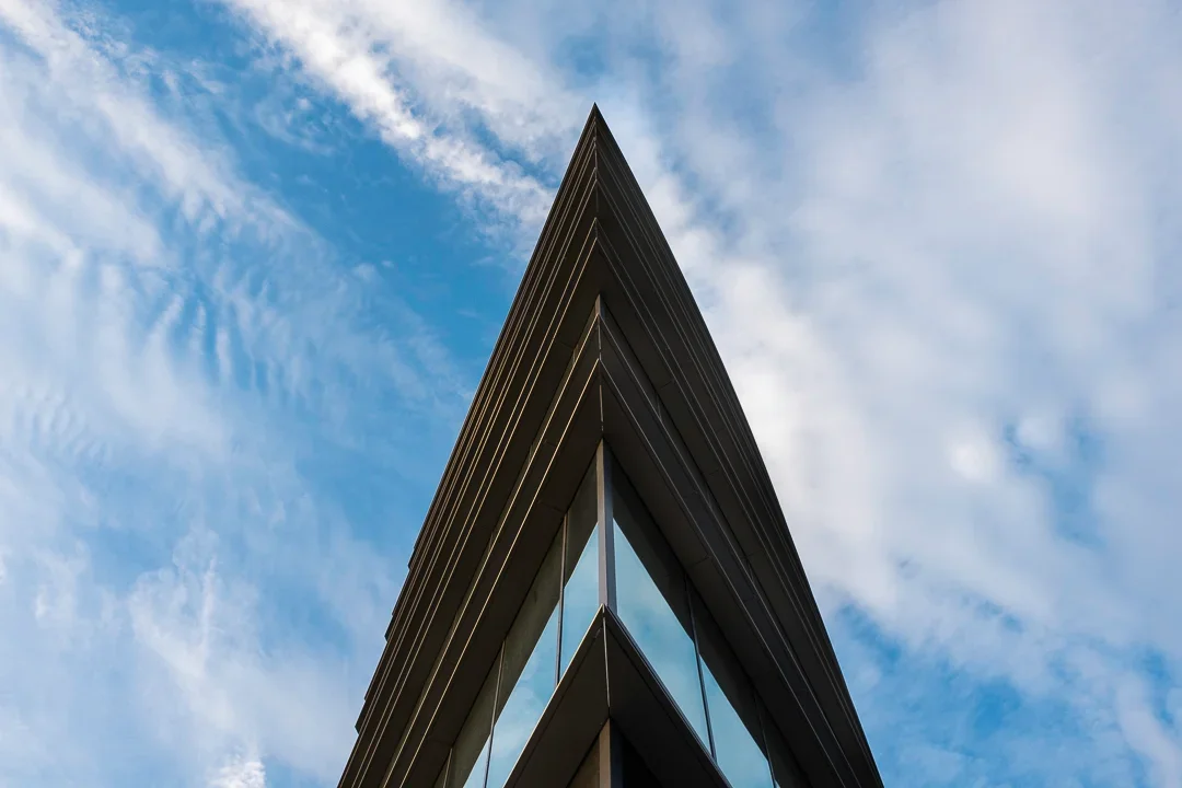 Close-up van de hoek van een modern gebouw met grote glasramen en een puntsgewijze top, tegen een blauwe lucht met wolken.