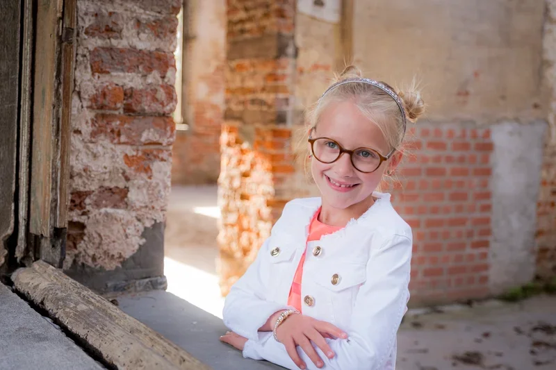 Lief meisje met bril, blond haar in een staart, glimlachend, in een witte jas, staand in een bouwval met bakstenen muren.