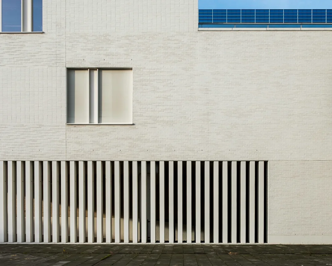 Witte muur met twee ramen en een decoratieve witte lattengevel aan de onderkant, op een straat met tegelstenen.