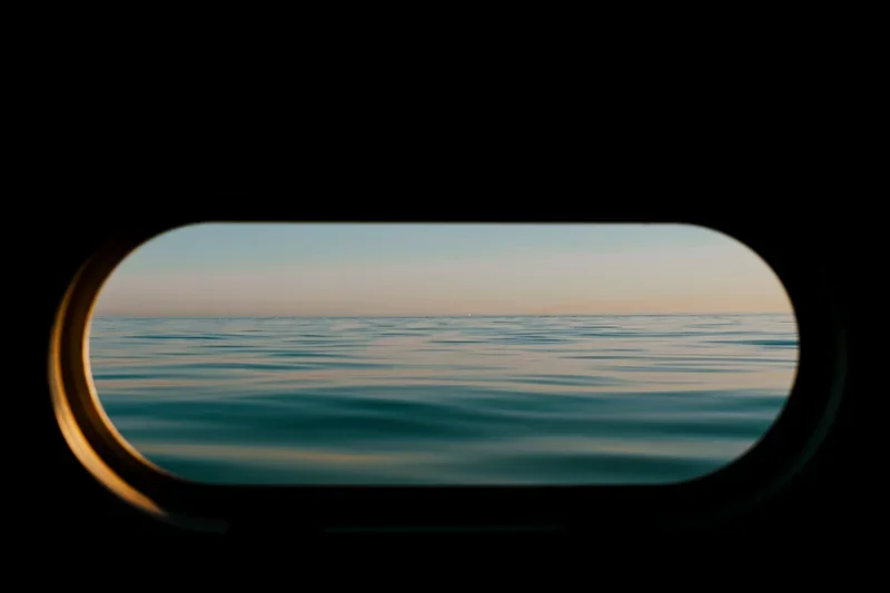 Uitzicht op zee of oceaan door een ronde raam van een schip of boot, met een rustige wateroppervlakte en een zonsondergang of zonsopgang op de horizon.