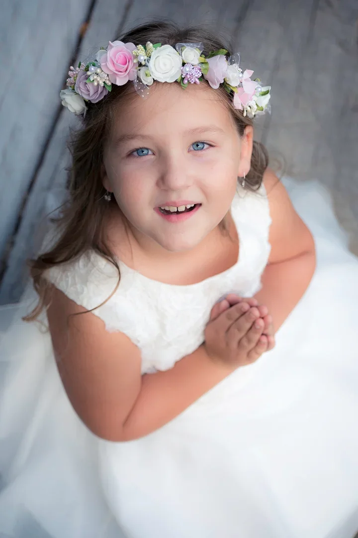 Een jong onschuldig meisje met een bloemenkrans op haar hoofd, witte jurk, glimlach, handen gevouwen, houten achtergrond.
