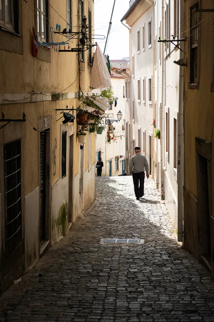 Een smal, geplaveid straatje tussen oude gebouwen met hangende waslijnen en planten, waarbij een man met een pet wegloopt.