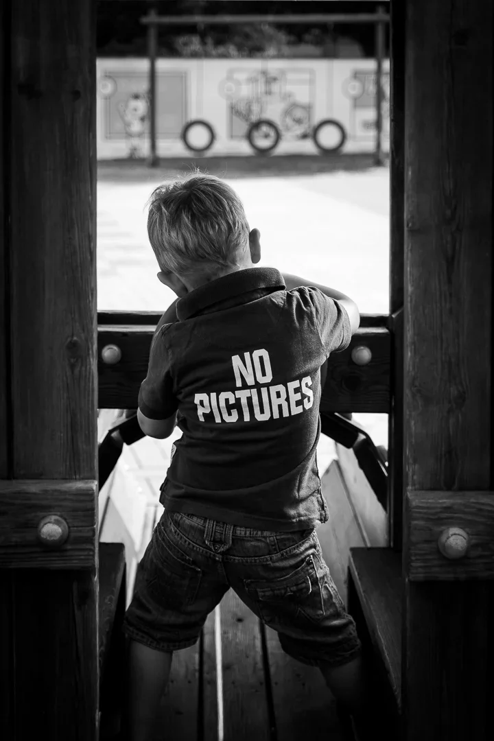 Een kind staat in een speeltoestel en kijkt uit op een schoolplein met een muur die graffiti en een schommel heeft.