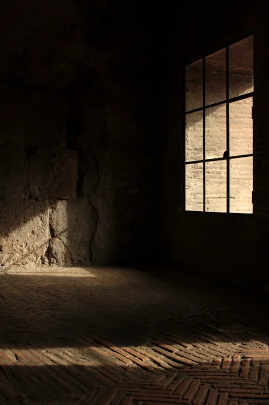 Een donkere gang of traphal met een stenen muur, een grote raam met smijdijzeren raamwerk, en een bakstenen vloer, licht een schaduw spel in Colosseum in Rome.