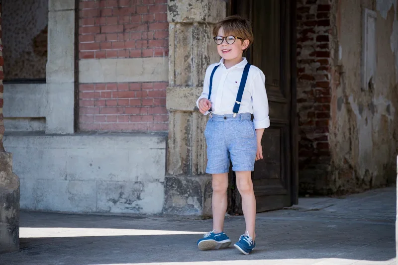 Jongen met bril, witte overhemd, korte blauwe broek, blauwe schoenen en een blauwe bretels, standend voor een oude stenen muur en een open deur in een stad of dorp.