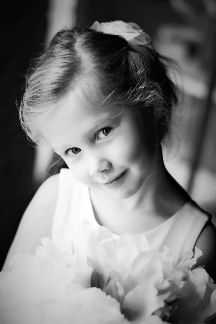 Zeer jonge meid met lichte haar in een paardenstaart met een grote lint, glimlachend en een boeket bloemen, in zwart-wit