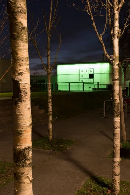 Een verlichte muur met de tekst 'MINUS ONE' in kruiswoordpatroon, zichtbaar vanaf een pad omringd door bomen, 's avonds of 's nachts.