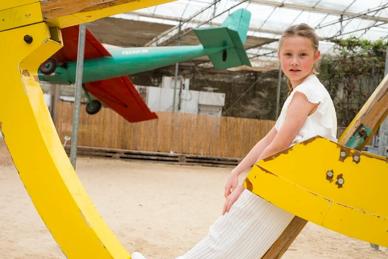 Een jonge meid zit op een houten schommel in een speeltuin met een stuk geel en een stuk groen speeltuig naast haar.