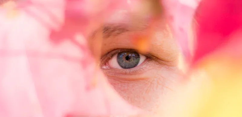 Close-up van een wit persoon met blauwe ogen, gedeeltelijk verborgen achter roze en gele bloemen.