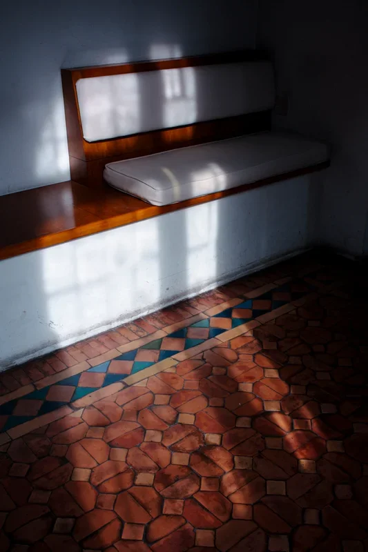 Een wit cushuys met houten rand en een matras op een witte muur in een kamer met een kleurrijke tegelvloer en schaduw van een raam op de muur.