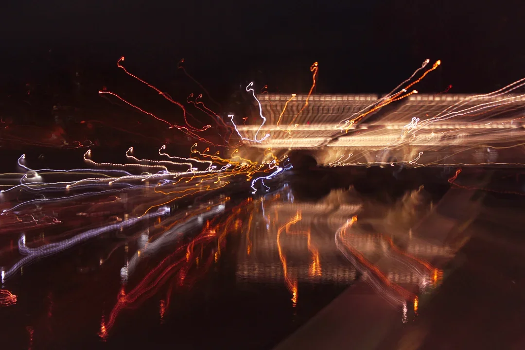 Lichtsporen van verlichte gebouwen of straatverlichting bij nacht, met reflecties op water of glad oppervlak, in een lang-expositie fotografiestijl.