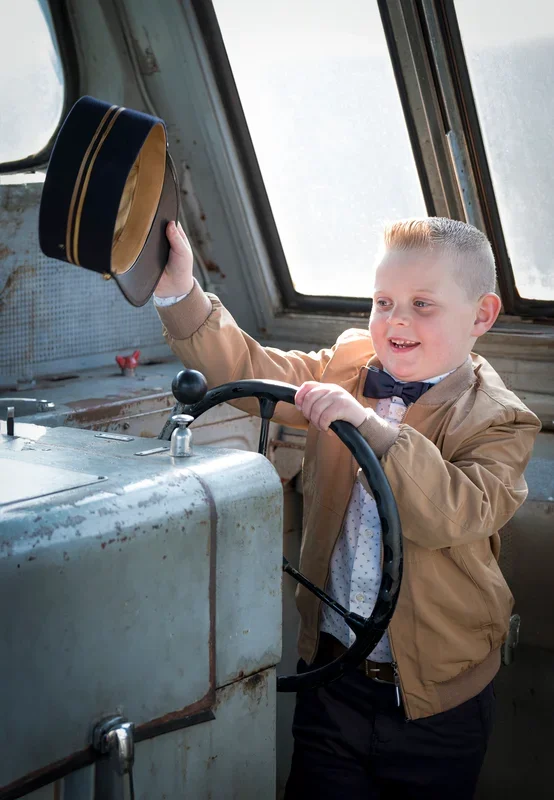 Een jongen met een blazer en een strikje in een locomotief van een oude stoomtrein, die een pet weglegt.