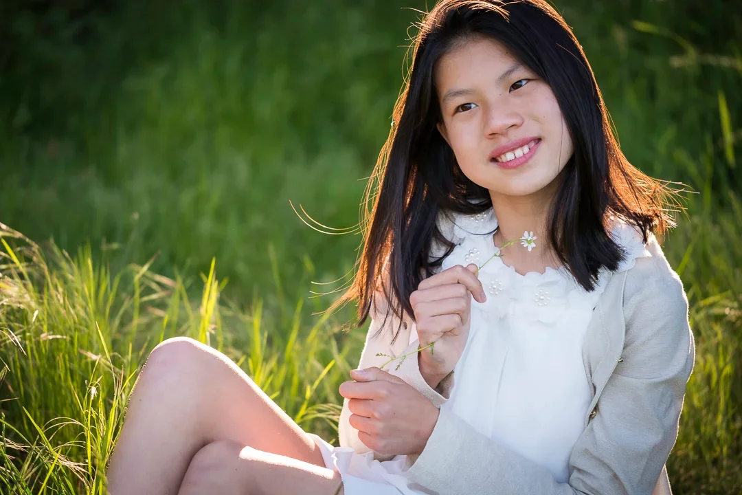 Een Aziatisch meisje met zwart haar zit buiten op gras, houdt een witte margriet in haar hand en kijkt naar rechts met een glimlach, omgeven door groen gras en bomen.