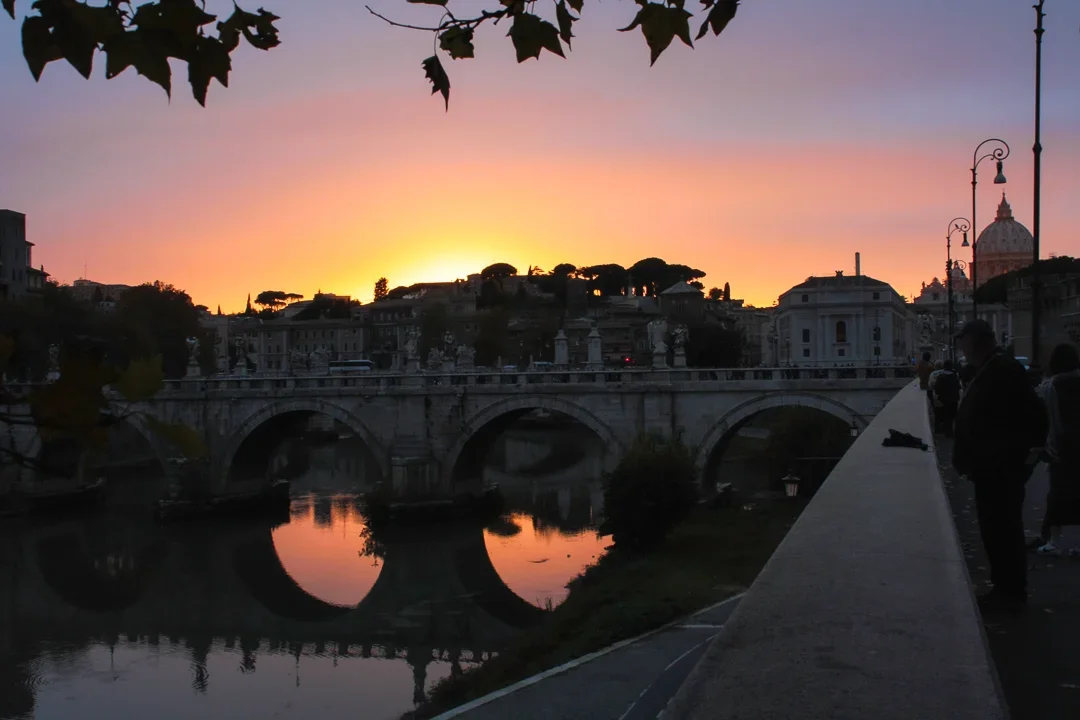 Gezicht op een brug over de rivier bij zonsondergang met silhouetten van mensen en gebouwen, en een gekleurde hemel.