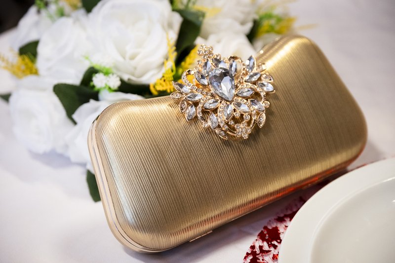 Gouden avondhandtas met een grote, kristallen bloemversiering, liggend naast een bloemenboeket.