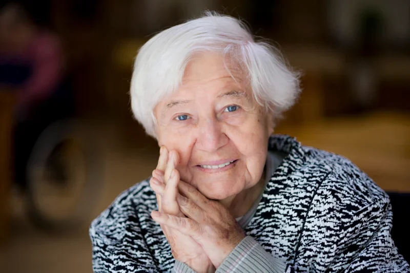 Een oudere vrouw met wit haar en blauwe ogen, glimlachend, rust haar hoofd op haar hand, zittend binnen.