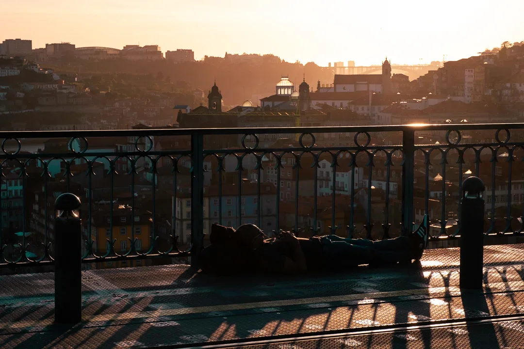 Persoon ligt op de grond bij een balkon of brug met uitzicht op een stad in de schemering/zonsondergang.