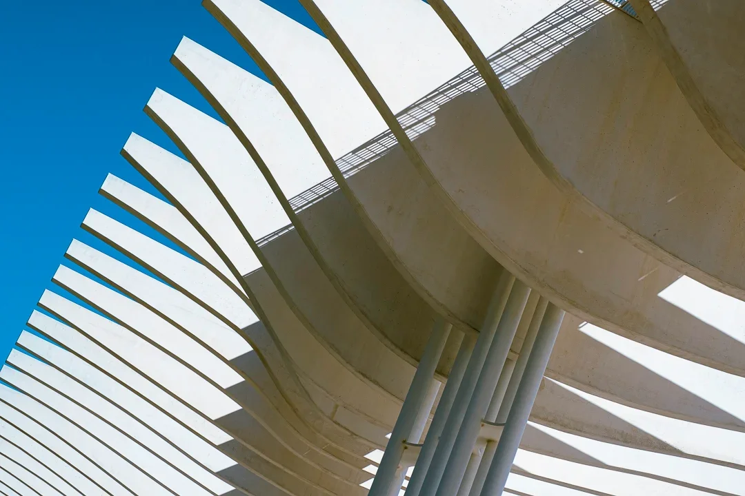 Een moderne architectonische structuur met grote, gebogen betonnen balken en verticale steunpijpen, met een blauwe hemel op de achtergrond.