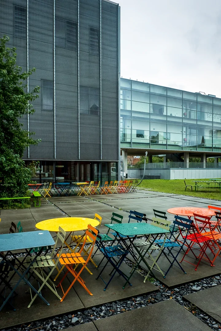 Kleurrijke tafels en stoelen op een terras buiten, met een modern kantoorpand op de achtergrond.