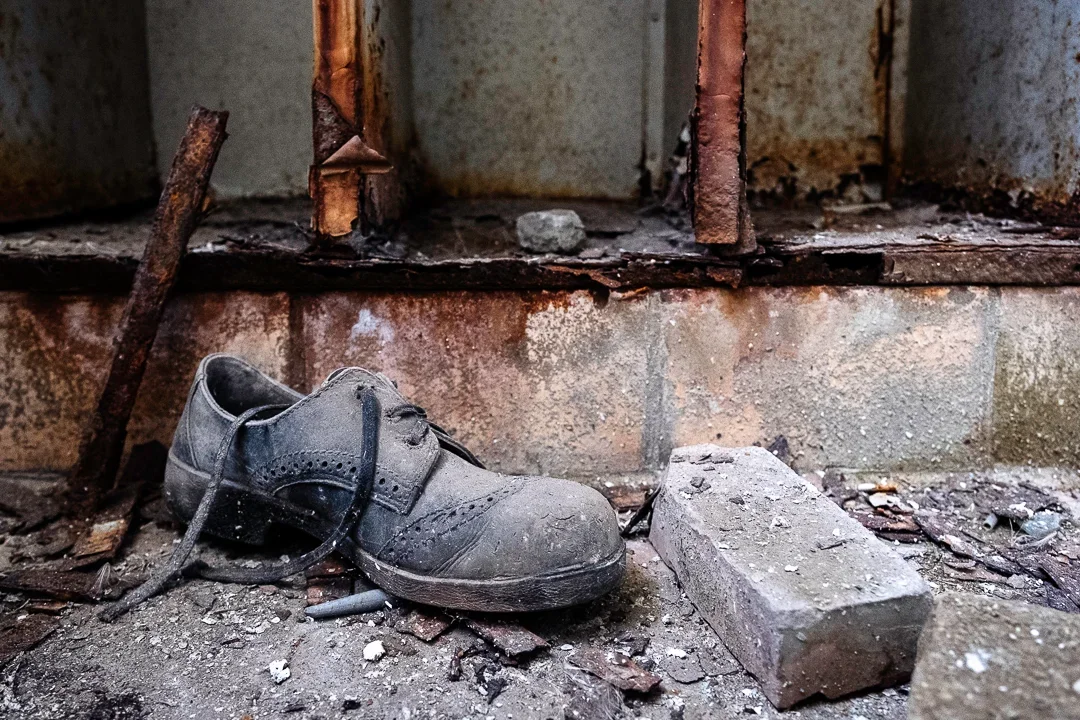 Een oude, versleten zwarte schoen ligt op de vuile, stoffige vloer van een verwaarloosde en roestige ruimte, met een bakstenen muur op de achtergrond.