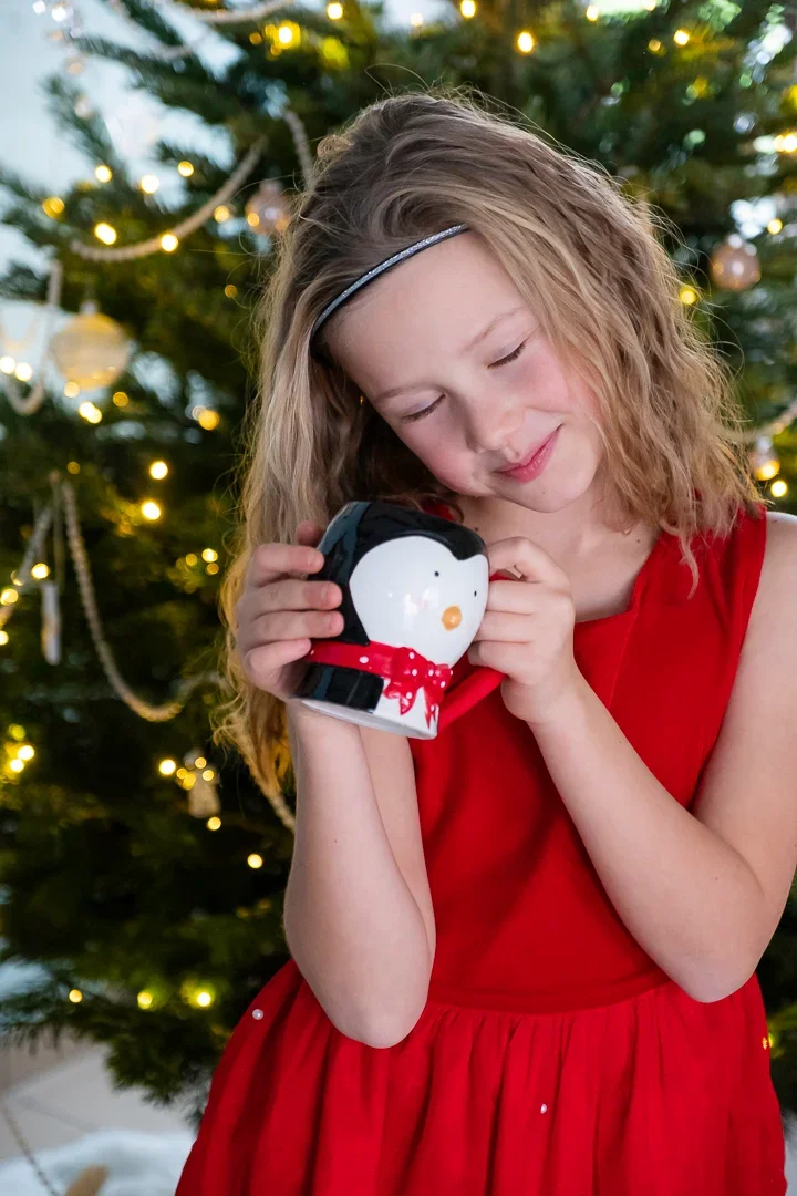 Een meisje in een rode jurk houdt een kerstkoekje met een pinguïn-kerstmok in haar handen, met een versierde kerstboom op de achtergrond.