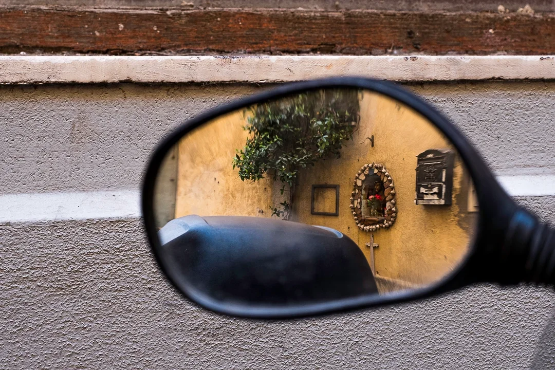 De foto toont een ronde spiegel die een muur weerspiegelt met een vegetatie en decoraties, waaronder een kruis en een adembescherming op een gele muur.