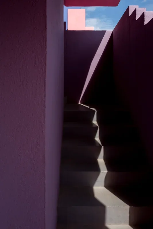 Foto van een steile trap met scherpe schaduwen, omringd door muren in pastelkleuren, onder een open hemel met enkele wolken.