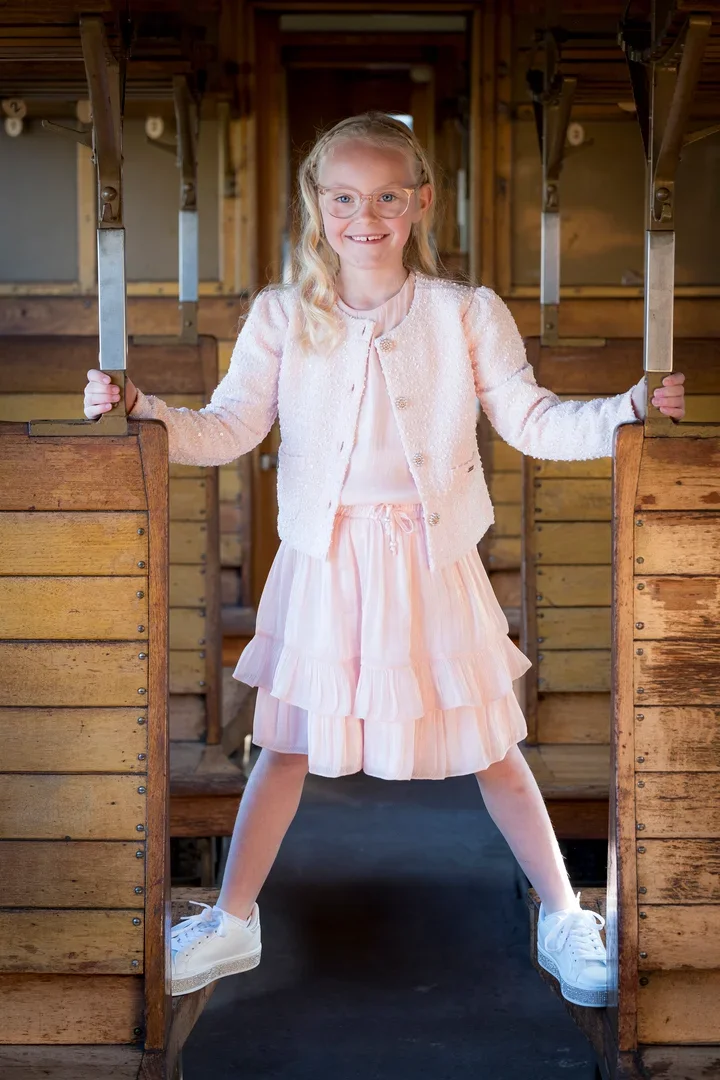 Vrolijke jong meisje met blonde haar en brillen, gekleed in een lichte roze jurk en een roze jas, staat stevig met haar voeten op een houten bankje in een oude houten wagon van een tre. Ze houdt de metalen handvatten vast en glimlacht naar de camera.