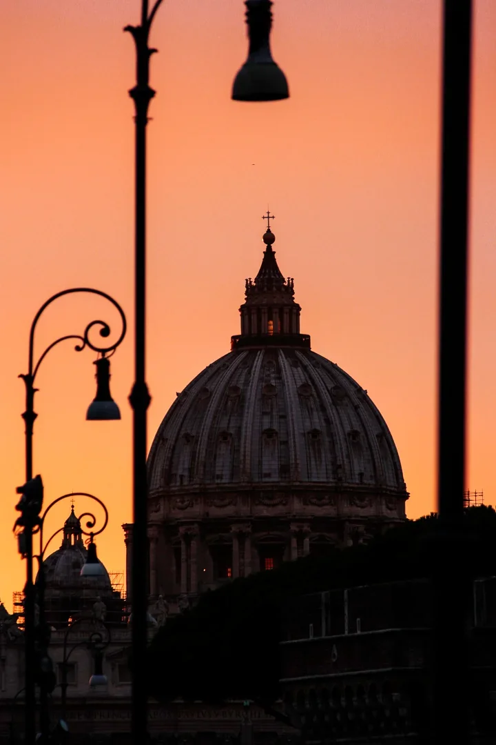 Uitzicht op de koepel van Sint-Pieter tijdens zonsondergang, met silhouetten van straatlantaarns op de voorgrond.