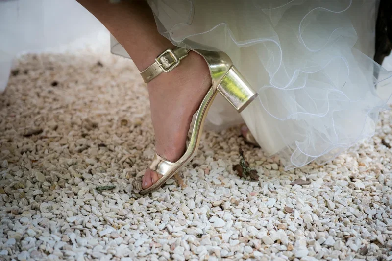 Een gouden schoen met een hoge hak die op een stenen ondergrond staat, bij een witte jurk.