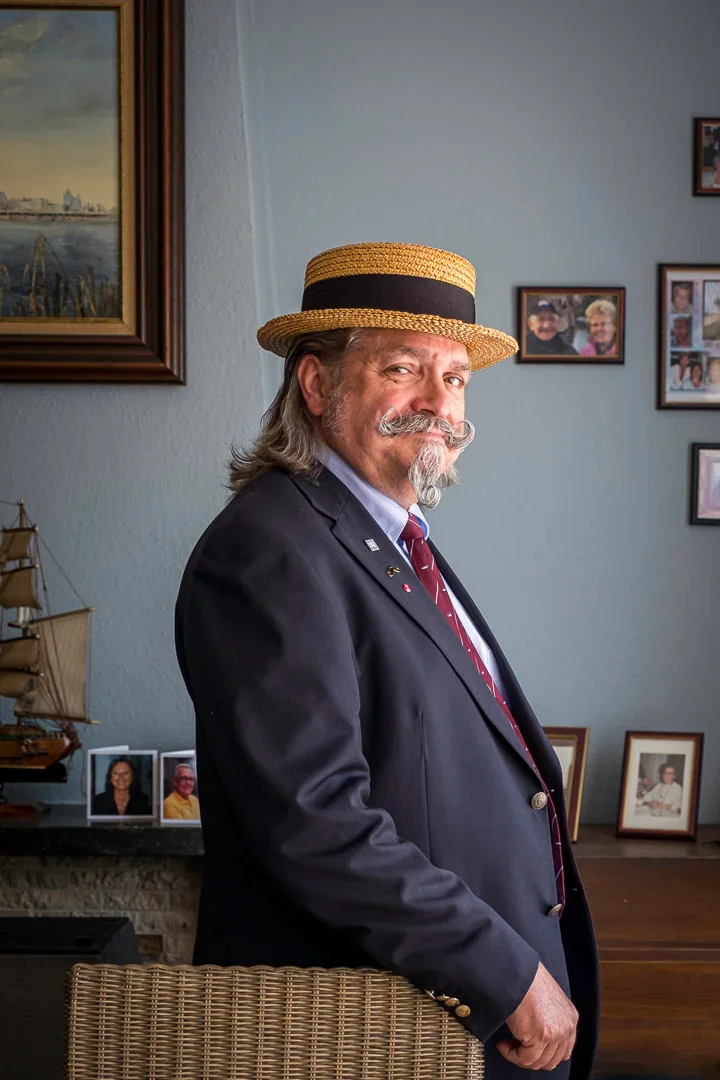 Man met grijze haar, snor en baard, gekleed in een donker pak, met een strooien hoed met zwarte band, staat binnen in een kamer met foto's en een model zeilschip op de achtergrond.