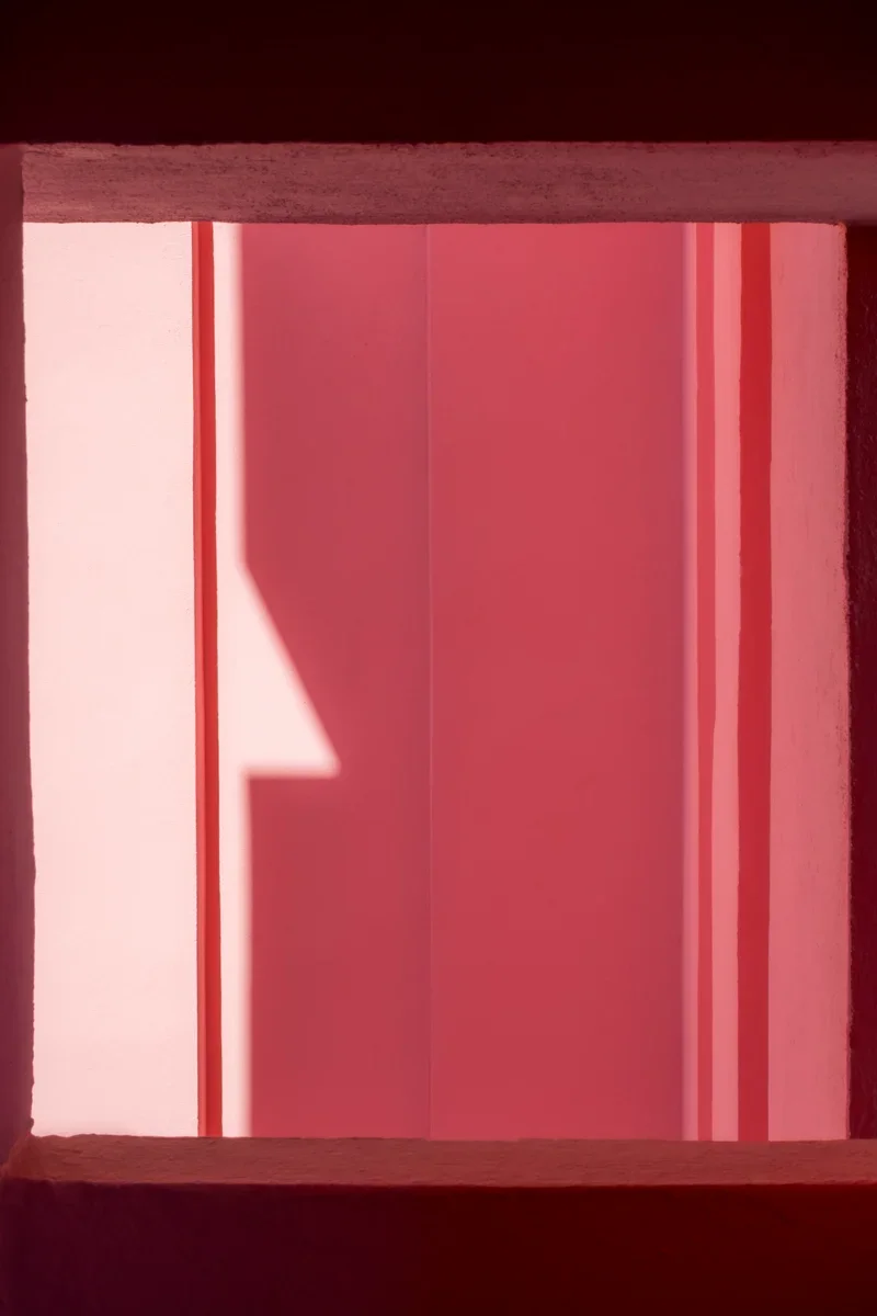 Een abstracte foto van een architecturaal patroon met roze en roodachtige tinten, weergegeven in een raam of opening met schaduwen en lichten.