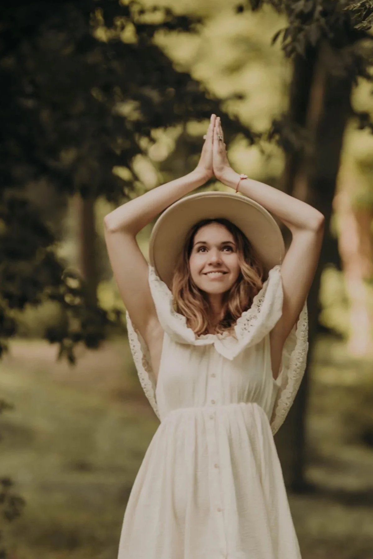 A young woman in a light-colored, sleeveless dress and wide-brimmed hat, standing outdoors with trees in the background, smiling and holding her hands in prayer above her head.