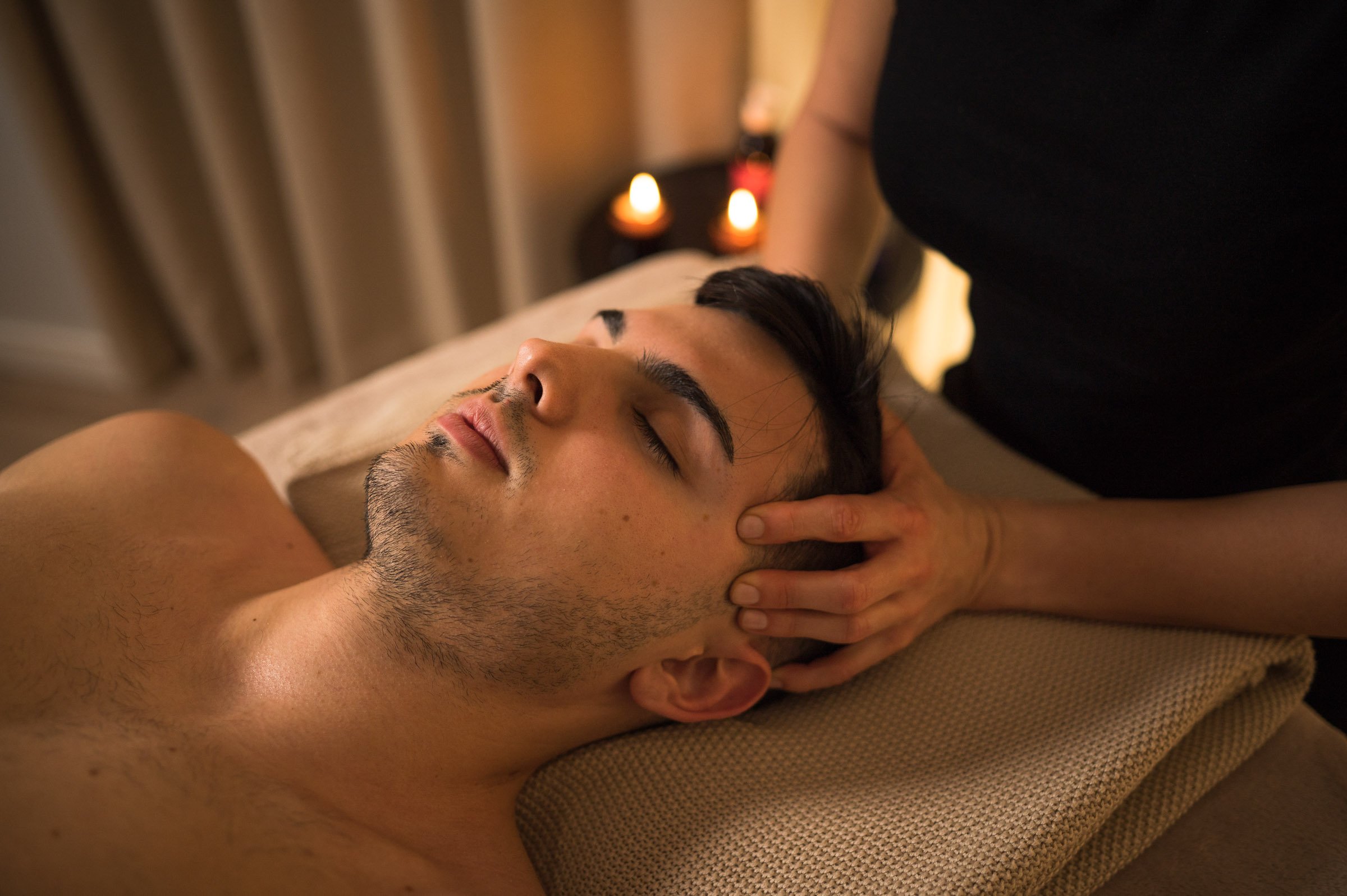 A man is lying on his stomach receiving a massage from a massage therapist. The man has his eyes closed, and the therapist is using her hands to massage his neck and shoulder area. There are candles in the background creating a relaxing ambiance.