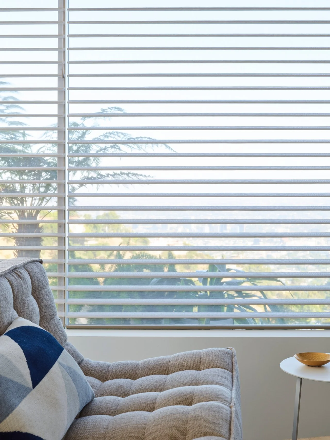 Living room with beige tufted sofa, blue and white geometric pillow, round white side table with a gold bowl, large window with white blinds, view of trees and blue sky outside.