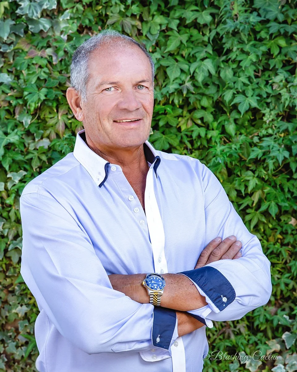 A man with short gray hair and blue eyes, wearing a white and navy button-up shirt with rolled-up sleeves, standing with arms crossed in front of a leafy green background.