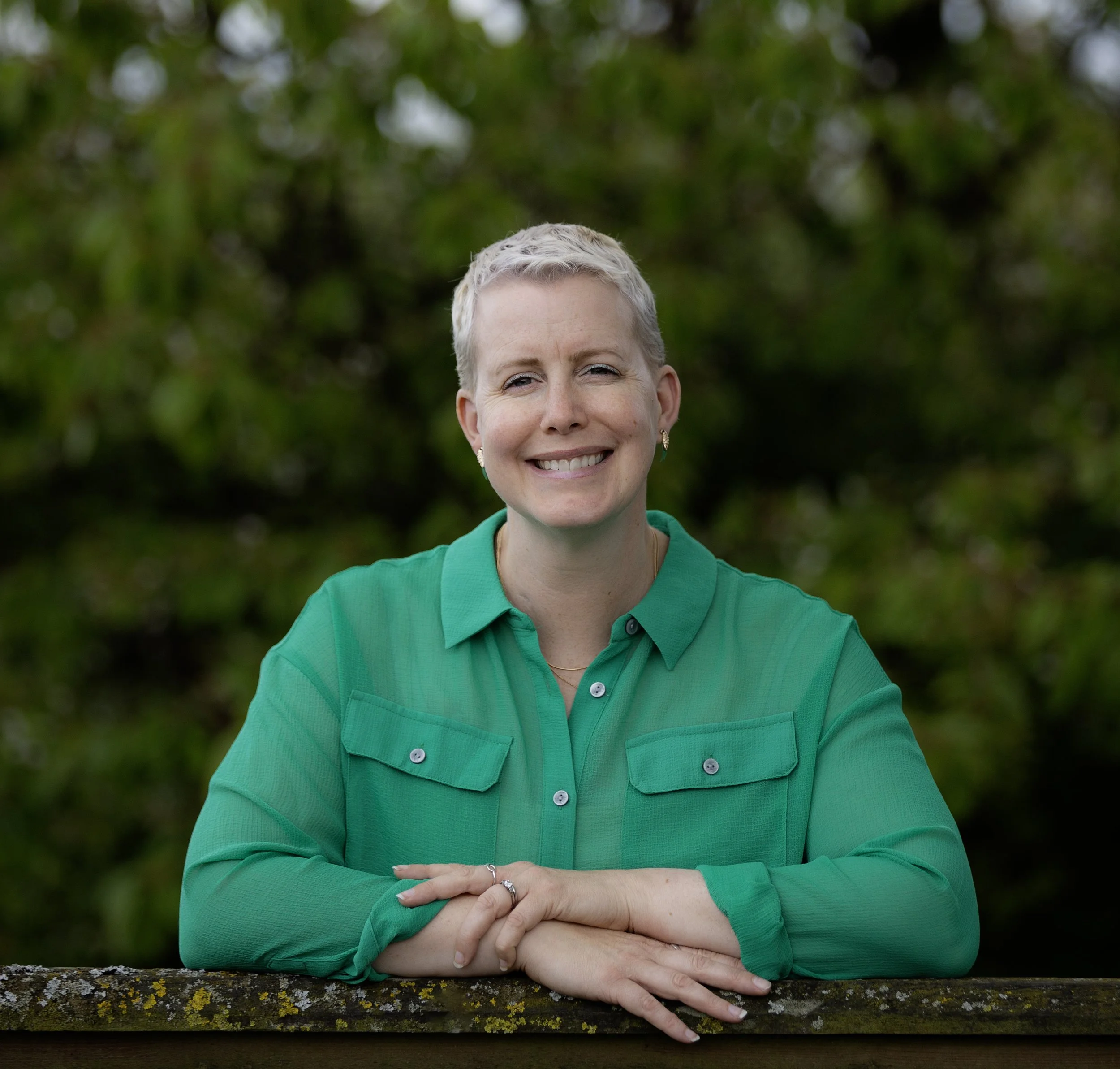Tara Ferguson, founder of Onward With You, wearing a green shirt, standing outdoors with a blurred green leafy background.