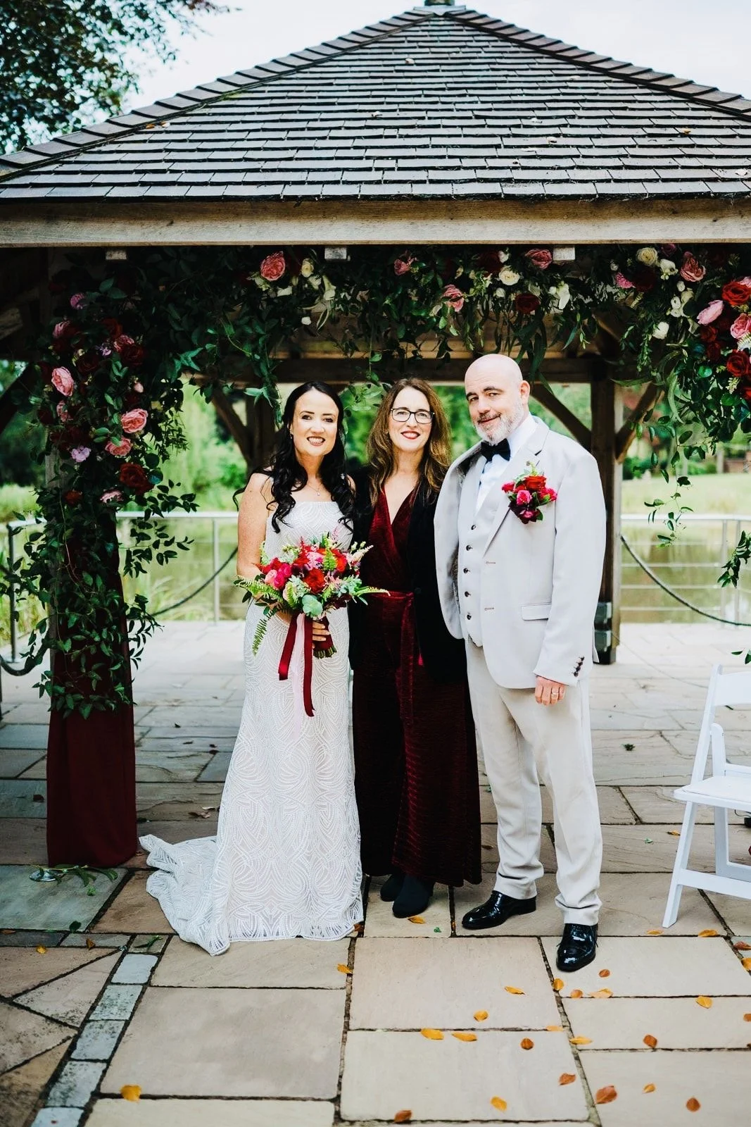 A wedding ceremony with three people standing under a wooden arch decorated with red and pink flowers. The bride is holding a bouquet and wearing a white dress, while the groom is in a white tuxedo with a black bow tie. The woman in the middle is wearing a dark burgundy dress. They are smiling and standing on a stone patio with a railing and greenery in the background.
