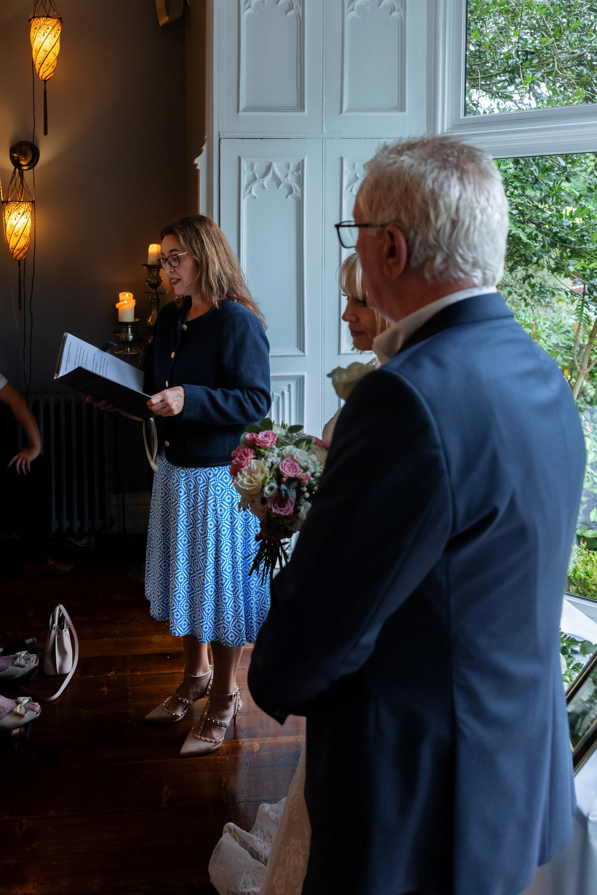 A woman is giving a speech or reading vows at a wedding ceremony in a decorated indoor space with candles and hanging lamps, while the bride and groom listen, with the groom holding a bouquet of flowers.