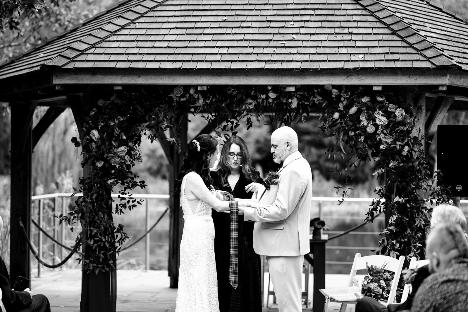 Black and white photo of a wedding ceremony at an outdoor gazebo, with a bride and groom holding hands and looking at each other, surrounded by friends and family.