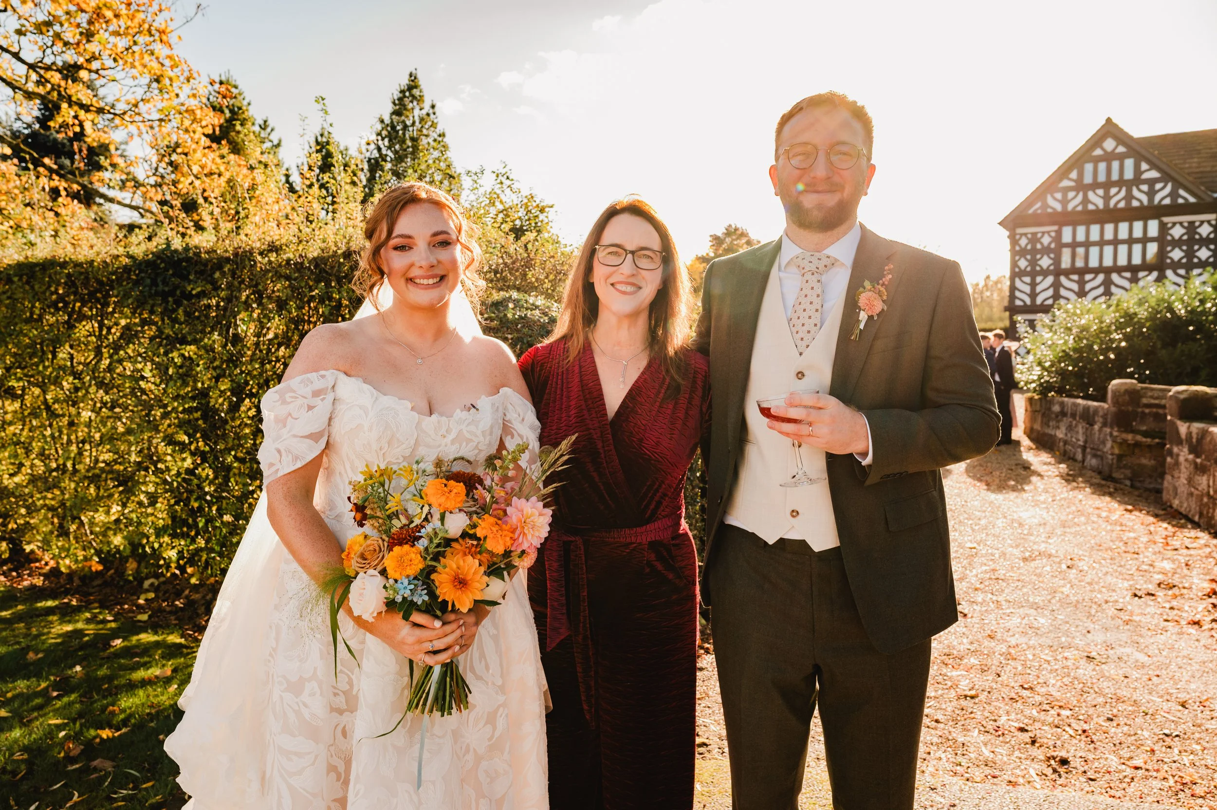 A bride in a white wedding dress holding a bouquet standing next to a woman in a red dress and a man in a suit holding a drink, outdoors during a sunny day.