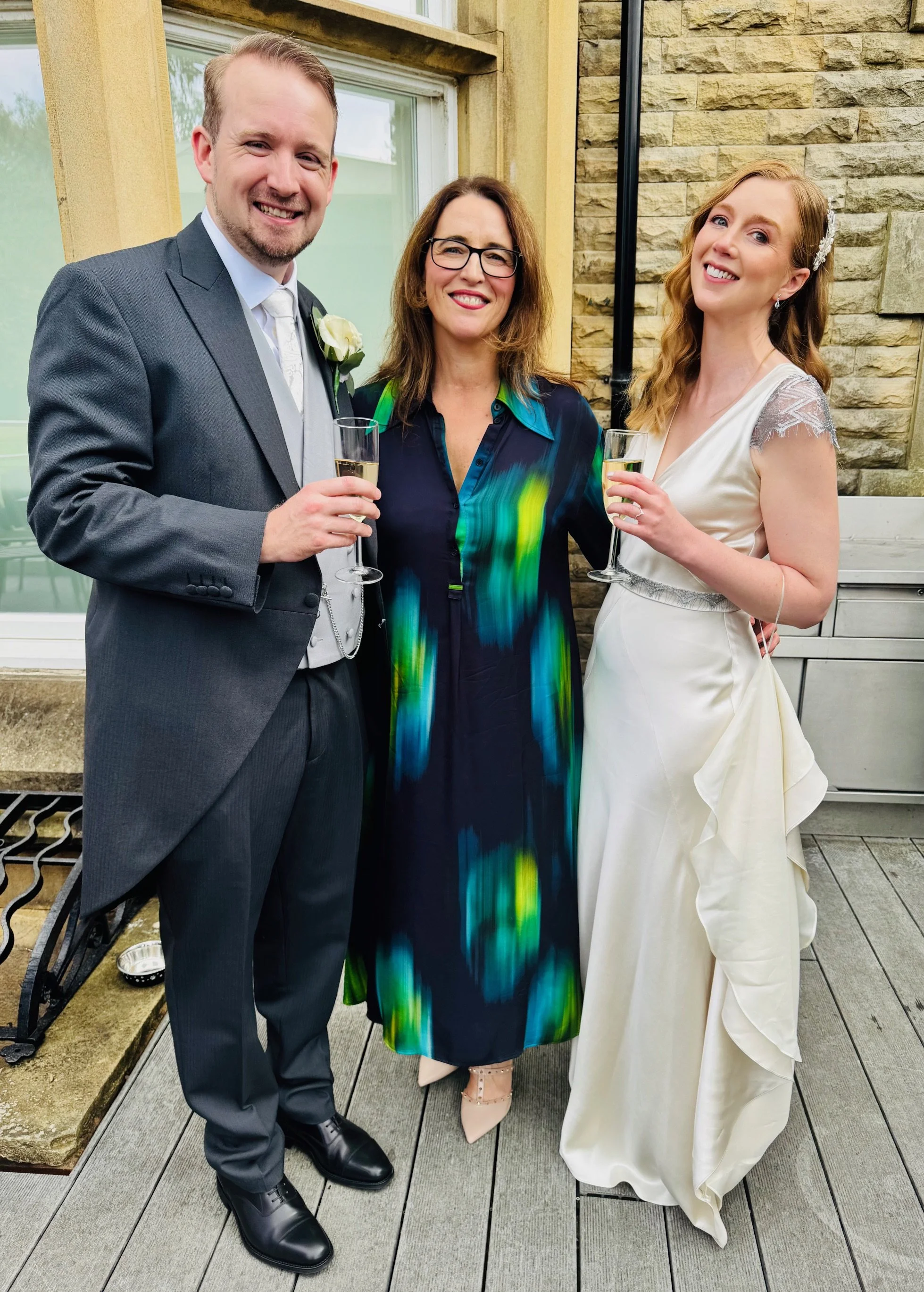 A man in a gray suit and a woman in a white dress are standing with another woman in a dark-colored dress that has bright, colorful patterns. They are all smiling and holding glasses of champagne. The setting appears to be outdoors, on a patio with a brick wall and a window in the background.